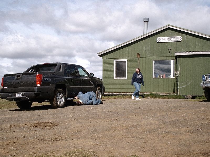 Black Chevrolet Avalanche pickup on dirt lot with person working underneath, green building behind.