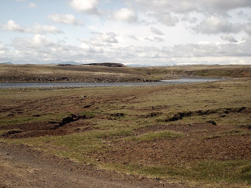Open grassland with a stream, low hills, and distant mountains under cloudy sky