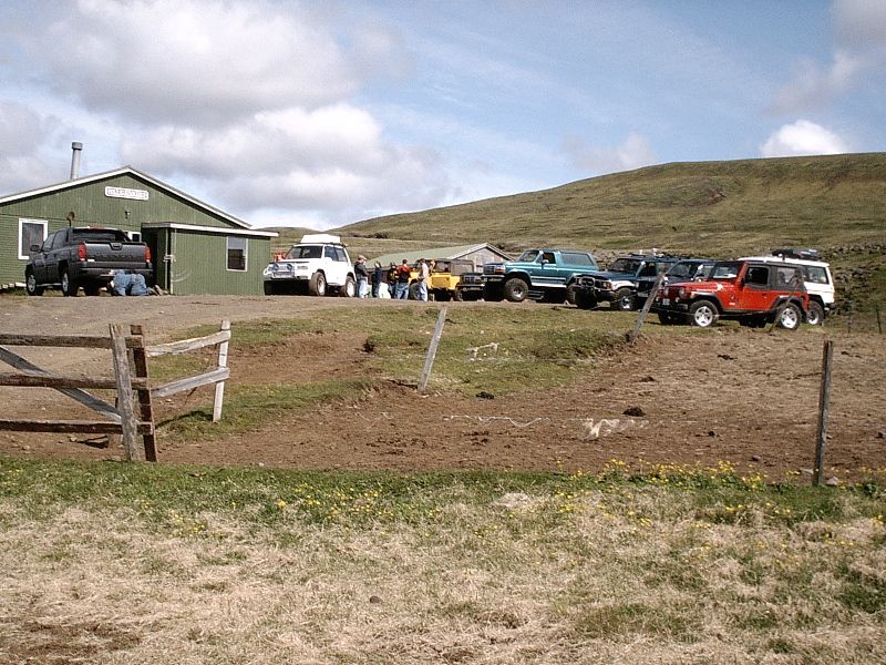 Off-road vehicles including a teal SUV and red Jeep parked in a grassy field by a green building