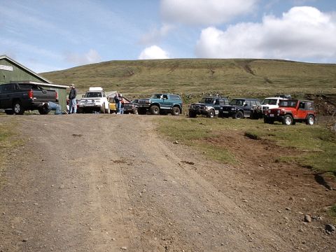 1996 Ford Bronco - A collection of off-road vehicles are gathered on a dirt path next to a green building.