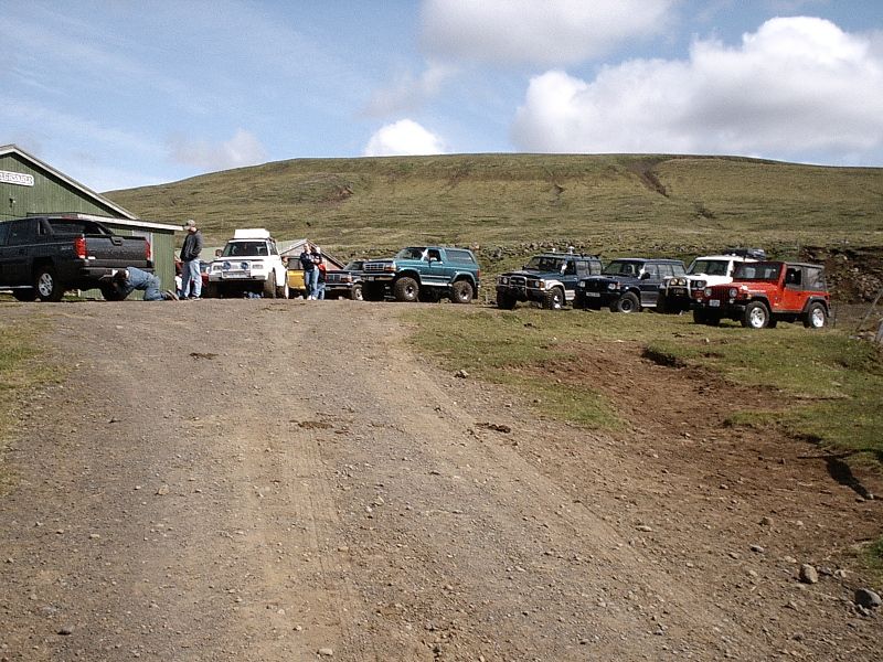 Row of lifted 4x4 SUVs and trucks parked on gravel below a grassy hill