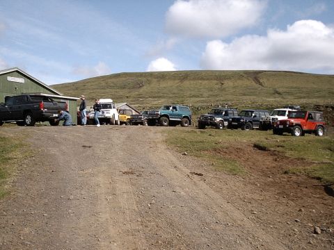 1996 Ford Bronco - A group of off-road vehicles are parked on a dirt road near a building on a sunny day.