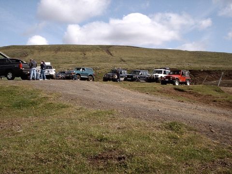 1996 Ford Bronco - A group of off-road vehicles are parked on a dirt path, with a grassy hill and cloudy sky overhead.