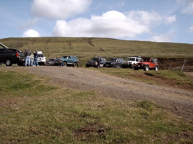 Row of parked off-road SUVs and a red Jeep on gravel in a grassy highland with people nearby.