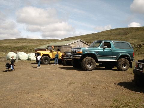 1996 Ford Bronco - Two off-road vehicles, a green Ford Bronco and a yellow vehicle, are parked outside a building on a sunny day.