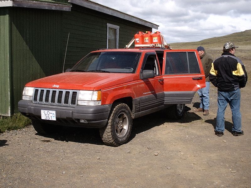 Red Jeep Grand Cherokee SUV parked by green building with roof-rack fuel cans and two men nearby