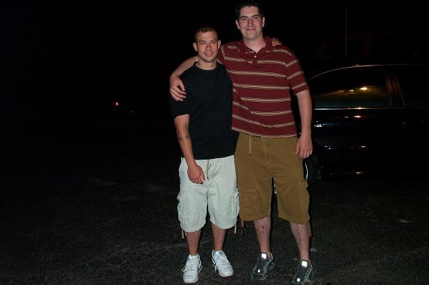 Two young men posing arm over shoulder at night beside a car