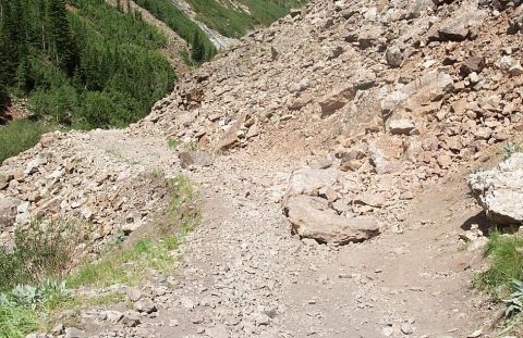 1990 Ford Bronco - A desolate mountain trail is covered in rocks and debris, with trees visible on the left slope.