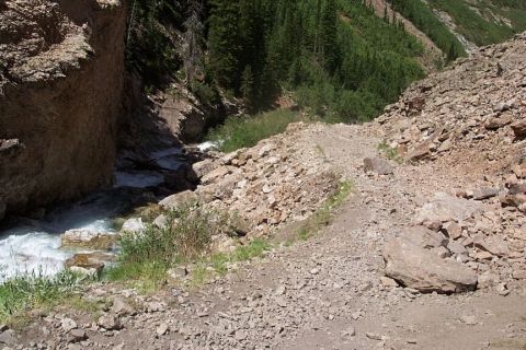 1990 Ford Bronco - A mountain trail winds alongside a fast-flowing river, surrounded by rocks and dense forest.