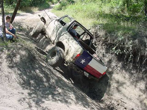 1978 Ford Bronco - A mud-covered lifted pickup truck with an open cab and a Texas flag painted on the tailgate climbs a steep dirt embankment on an off-road t…