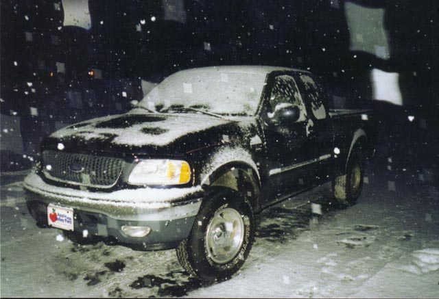 Dark Ford F-150 pickup covered in snow at night, front-left view