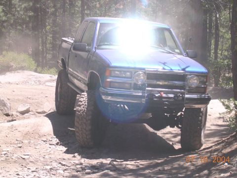 1994 Ford Bronco - A lifted blue pickup truck navigates a dusty, rocky trail through a wooded area.