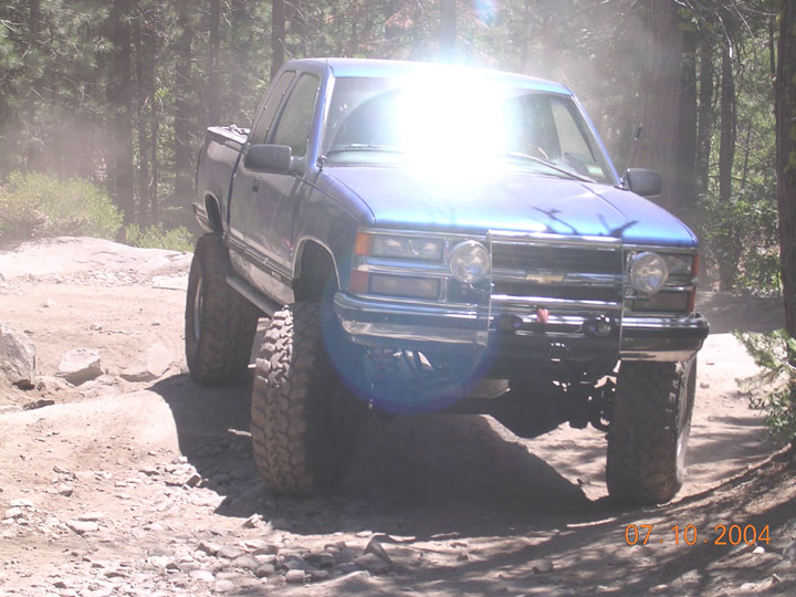 Blue pickup truck with large tires on a dirt trail, sun glare on windshield. Date stamp reads 07 10 2004