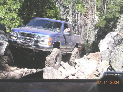 1994 Ford Bronco - A blue pickup truck with oversized tires is shown off-roading on a rocky path.