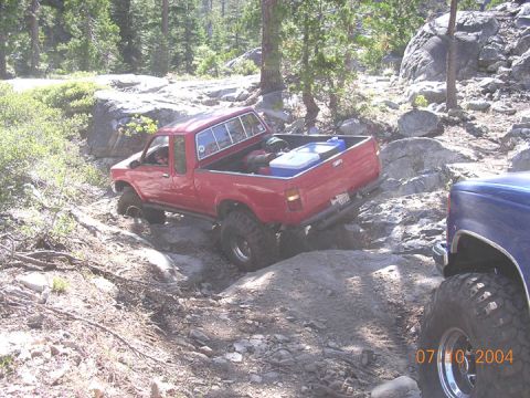 1994 Ford Bronco - A red pickup truck is shown off-roading on a rocky trail, with another vehicle partially visible in the foreground.