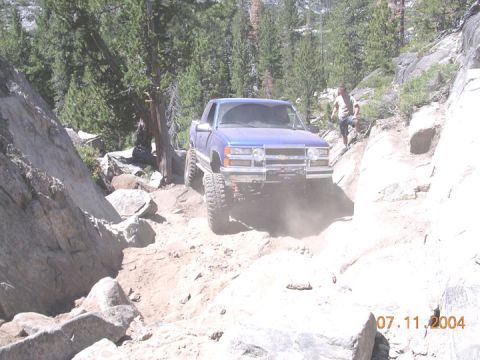 1994 Ford Bronco - A blue pickup truck navigates a challenging rocky off-road path on a sunny day.