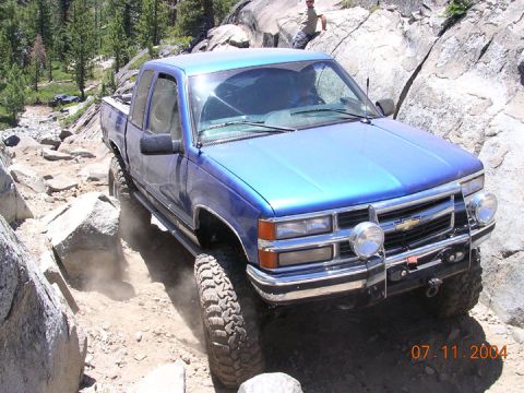 1994 Ford Bronco - A blue pickup truck is shown off-roading on a rocky path, with another vehicle visible in the background.