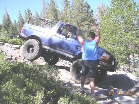 1994 Ford Bronco - A man cheers as a modified blue SUV navigates a rocky trail on a sunny day.