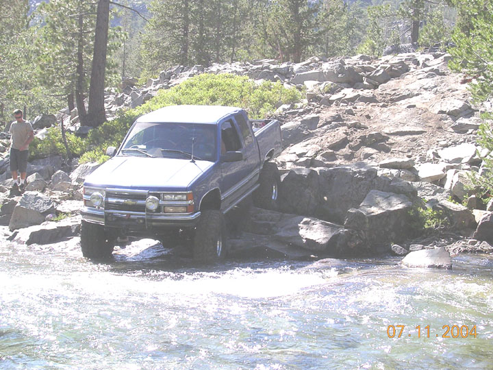 Lifted blue extended-cab pickup crawling off granite rocks into a rushing river