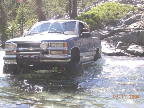 1994 Ford Bronco - A lifted blue pickup truck navigates a shallow, rocky stream on a sunny day.