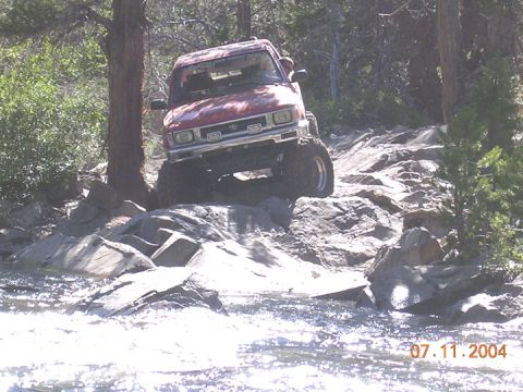 1994 Ford Bronco - Toyota pickup truck tackling a challenging rocky trail with water in the foreground.