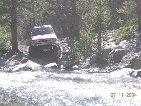 1994 Ford Bronco - A red off-road vehicle drives through a shallow, rocky riverbed on a sunny day.