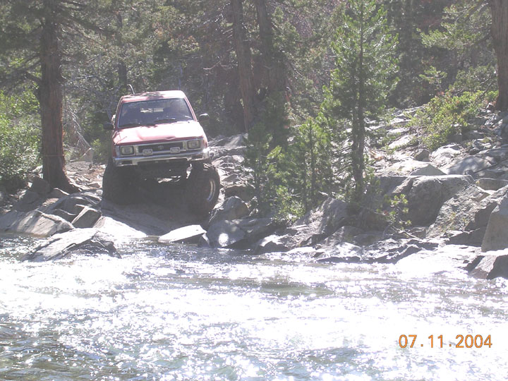 Red SUV navigating a rocky stream with trees and boulders around