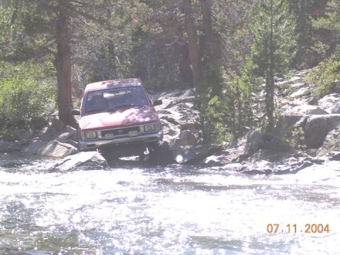 1994 Ford Bronco - A red SUV is shown off-roading through a rocky stream with trees in the background.