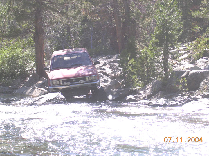 Red SUV navigating a rocky stream, front view, with trees and rocks surrounding
