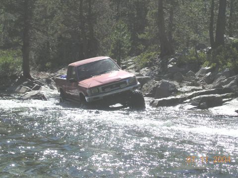 1994 Ford Bronco - A red pickup truck, likely from 1994 based on its body style, drives through a rocky stream on a sunny day.
