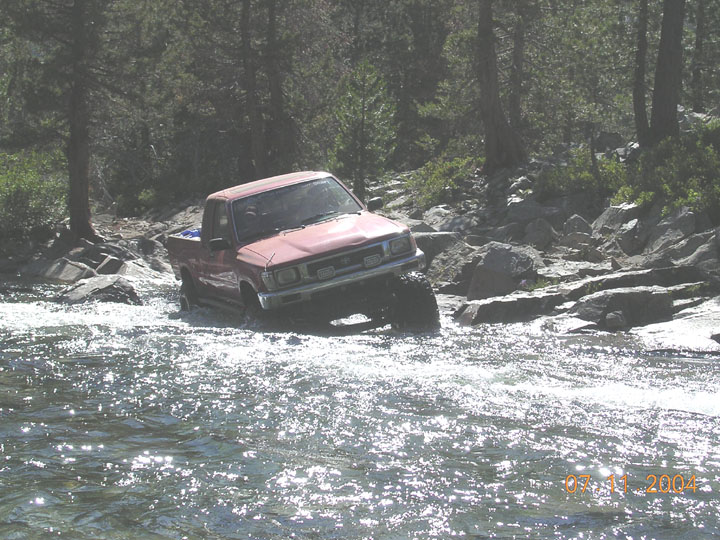 Red compact 4x4 pickup fording a rocky sunlit river in the pines, 2004