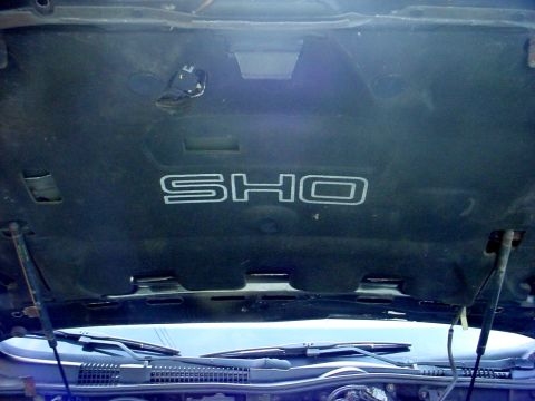 1989 Ford Taurus - The underside of a car hood displays the 'SHO' emblem, with visible hood supports and windshield wipers below.