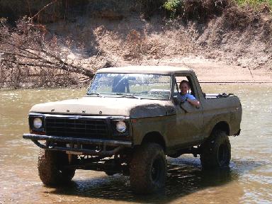 1978 Ford Bronco - Muddy tan topless Ford Bronco with FORD hood lettering sitting axle-deep in a shallow river, shot from the 3/4 front with a driver leaning…