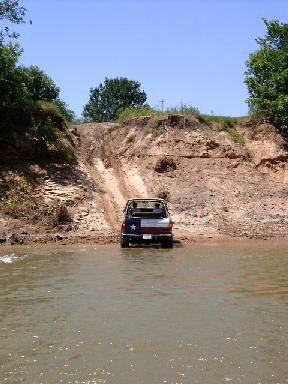 1978 Ford Bronco - A 4x4 with a Texas-flag-painted tailgate sits at the water's edge of a muddy river after descending a steep eroded dirt bank, photographed…