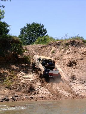 1978 Ford Bronco - A light-colored full-size Bronco-style 4x4 descends a steep, eroded dirt bank toward a muddy river, kicking up dust as it drops. Shot from…