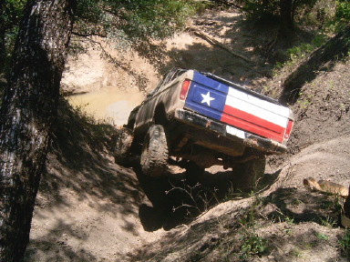 1978 Ford Bronco - Rear view of a mud-covered full-size Ford Bronco flexing through a rutted, muddy trail gully in the woods, its tailgate painted as a Texas…