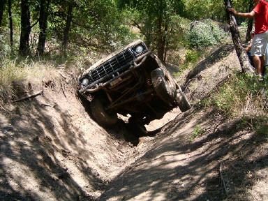 1978 Ford Bronco - A muddy full-size Ford Bronco tips hard onto one side as it climbs out of a steep-walled dirt gully on a wooded trail, front end pointed up…