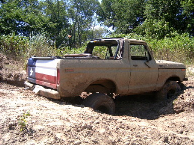 1978 Ford Bronco - A topless full-size Ford Bronco with a Texas-flag-painted tailgate sits buried to the axles in a churned mud pit, mud packed onto the body…