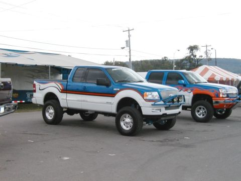 1994 Ford Bronco - Two custom lifted pickup trucks are parked side-by-side, with the truck in the foreground a white and blue paint scheme and custom decals.