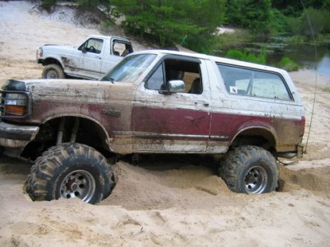 1993 Ford Bronco - A white and maroon two-tone Ford Bronco SUV with a lift kit and mud tires sits stuck in deep sand, its body caked in dried mud, shown in si…