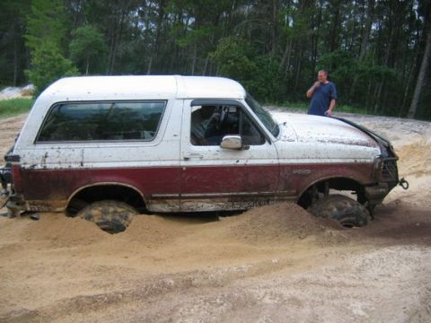 1993 Ford Bronco - Side view of a mud-splattered white-over-maroon two-door Ford Bronco buried to the axles in loose sand, with mud-terrain tires dug into rut…