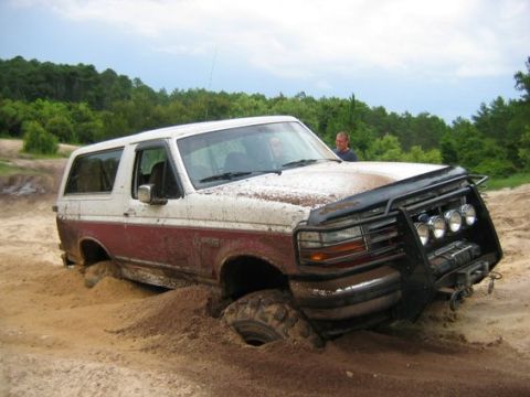 1993 Ford Bronco - A mud-spattered burgundy and white full-size Bronco buried to the axles in a sand pit, leaning nose-down with two people standing behind it…