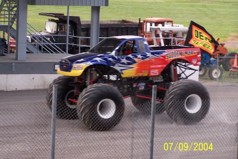 1979 Ford Bronco - A monster truck with patriotic graphics is seen in motion, with other vehicles visible behind a fence.