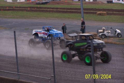 1979 Ford Bronco - Monster trucks compete on a dusty track, kicking up dirt as they race past the fence.