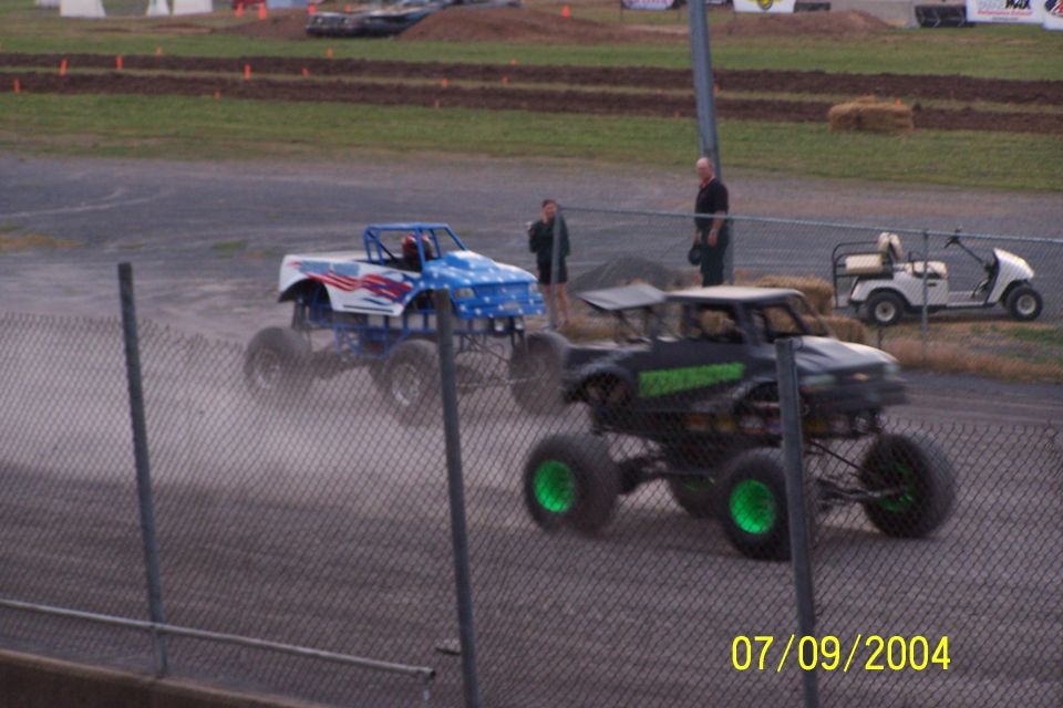 Two monster trucks race on a dirt track, one with patriotic paint, the other black with green wheels. A golf cart is parked nearby