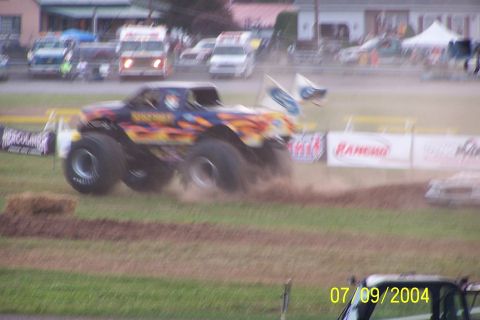 1979 Ford Bronco - A monster truck with flame graphics kicks up dirt as it drives across a track, with other vehicles blurred in the background.