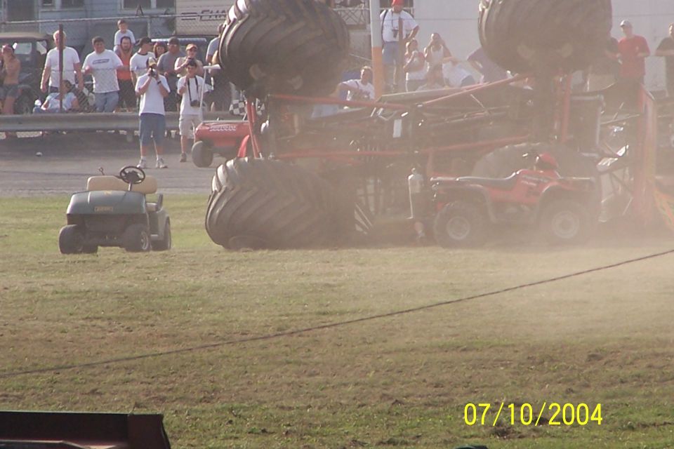 Monster truck on its side with an ATV nearby and a golf cart in the foreground. Spectators watch from a raised platform