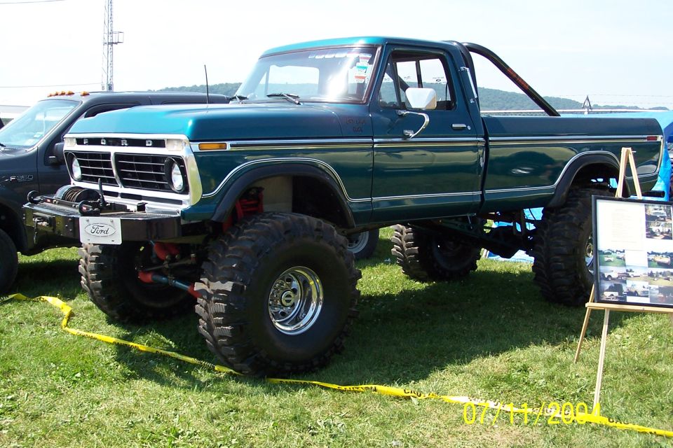 Lifted teal Ford pickup truck with large tires and a roll bar. Another dark pickup is parked nearby