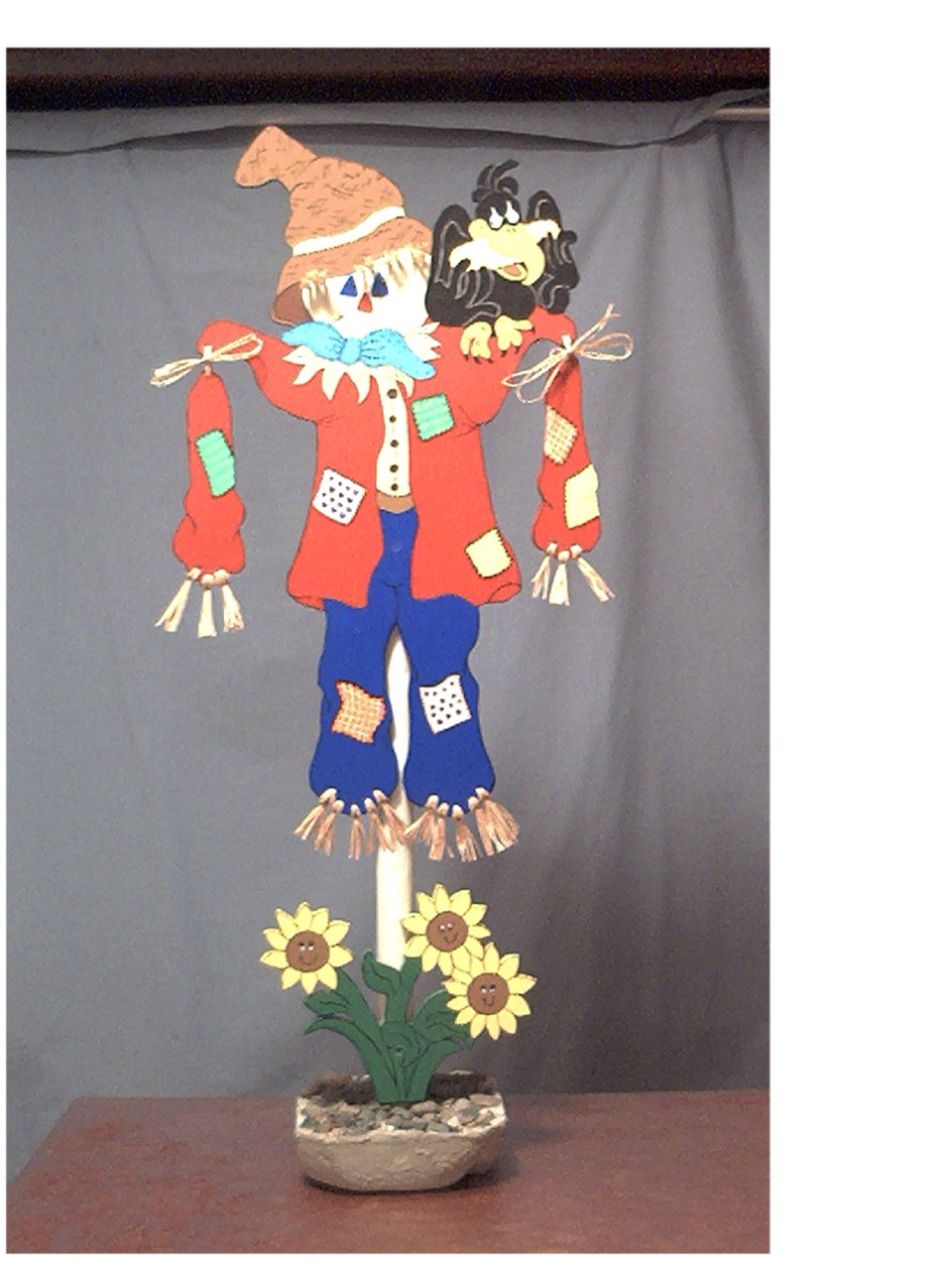Cardboard scarecrow decoration with a crow on its shoulder, wearing a hat and patched clothes, with sunflowers at its base