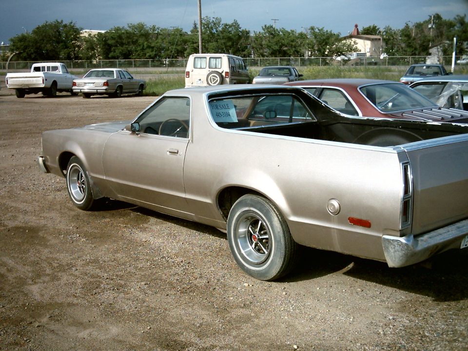 Beige Ford Ranchero pickup, rear three-quarter view, with a For Sale sign in the window on a gravel lot